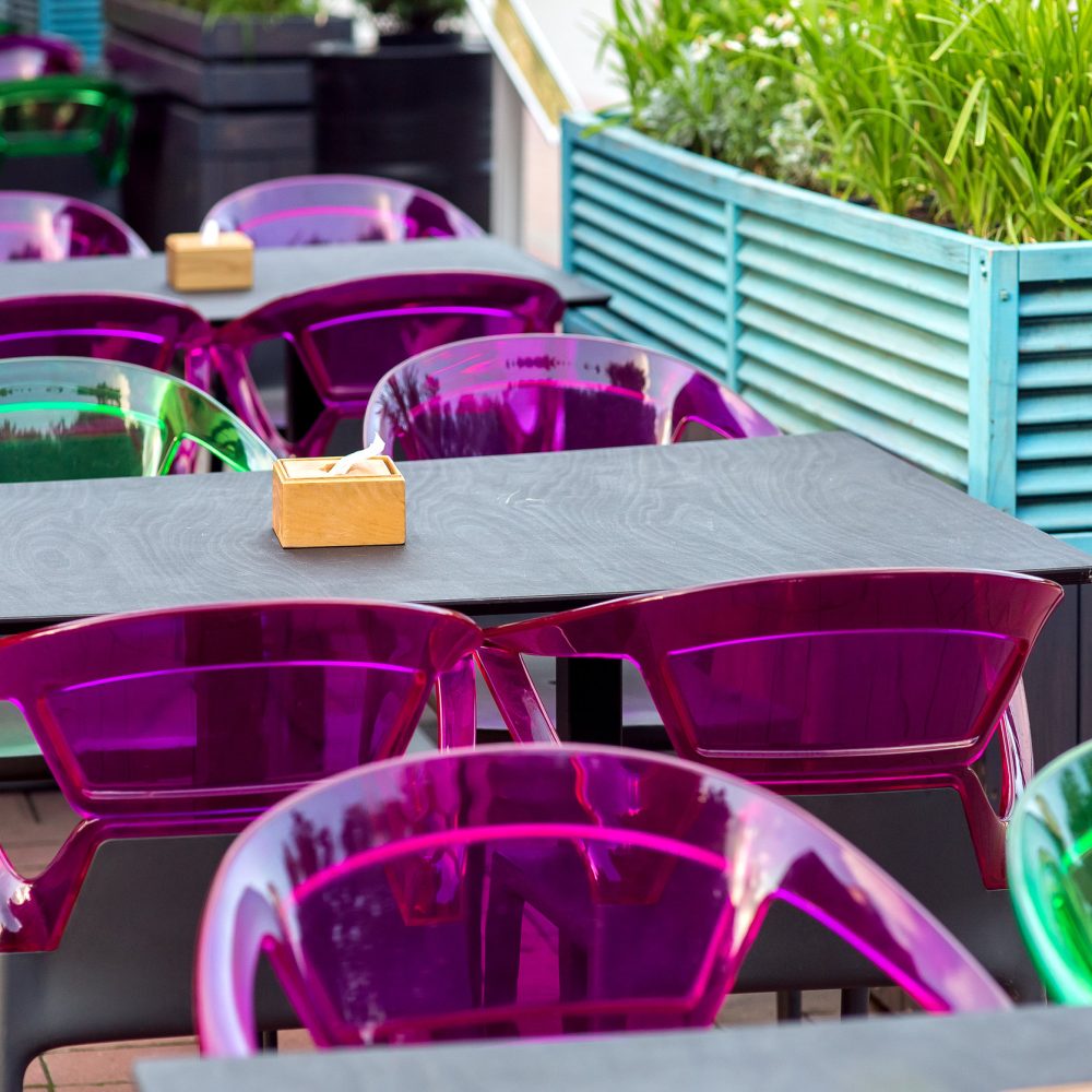 a wooden table top with a napkin holder and plastic multi-colored chairs on the terrace of a street cafe with free seats, nobody.